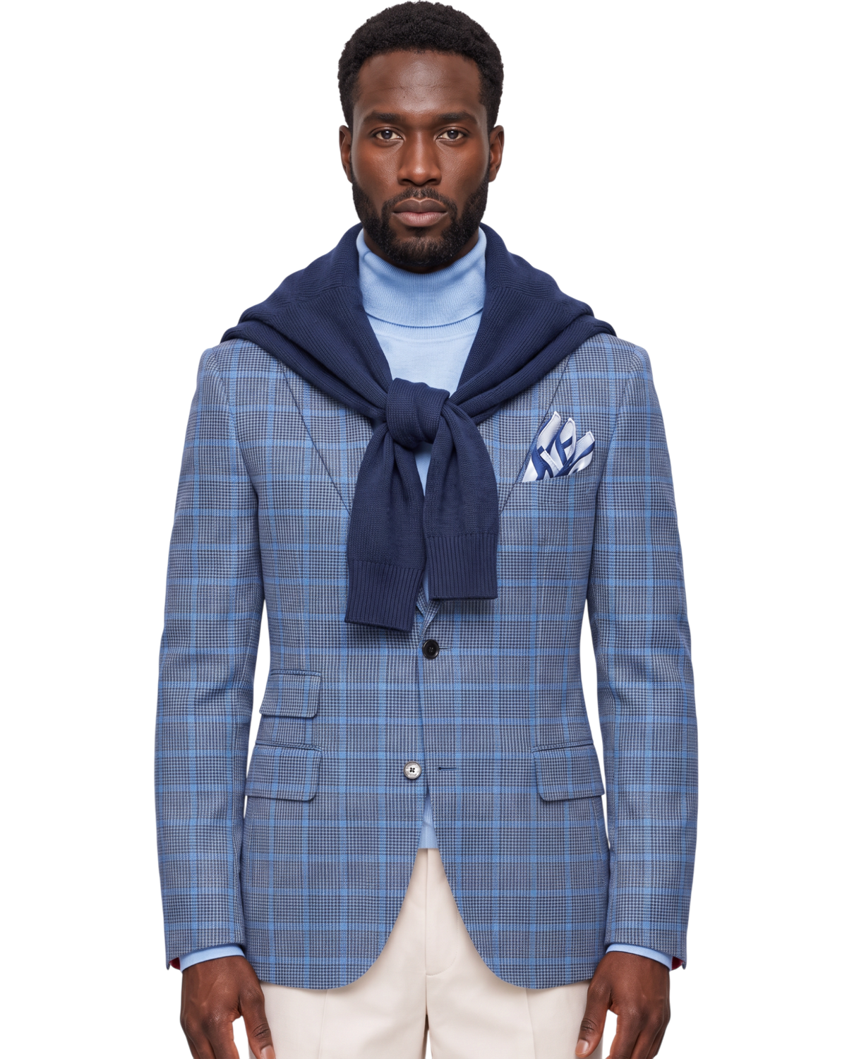 A man modeling a blue and gray plaid blazer over a light blue turtleneck sweater, styled with a navy blue knit hood draped over his shoulders, cream trousers, and a matching patterned pocket square, against a plain white background.