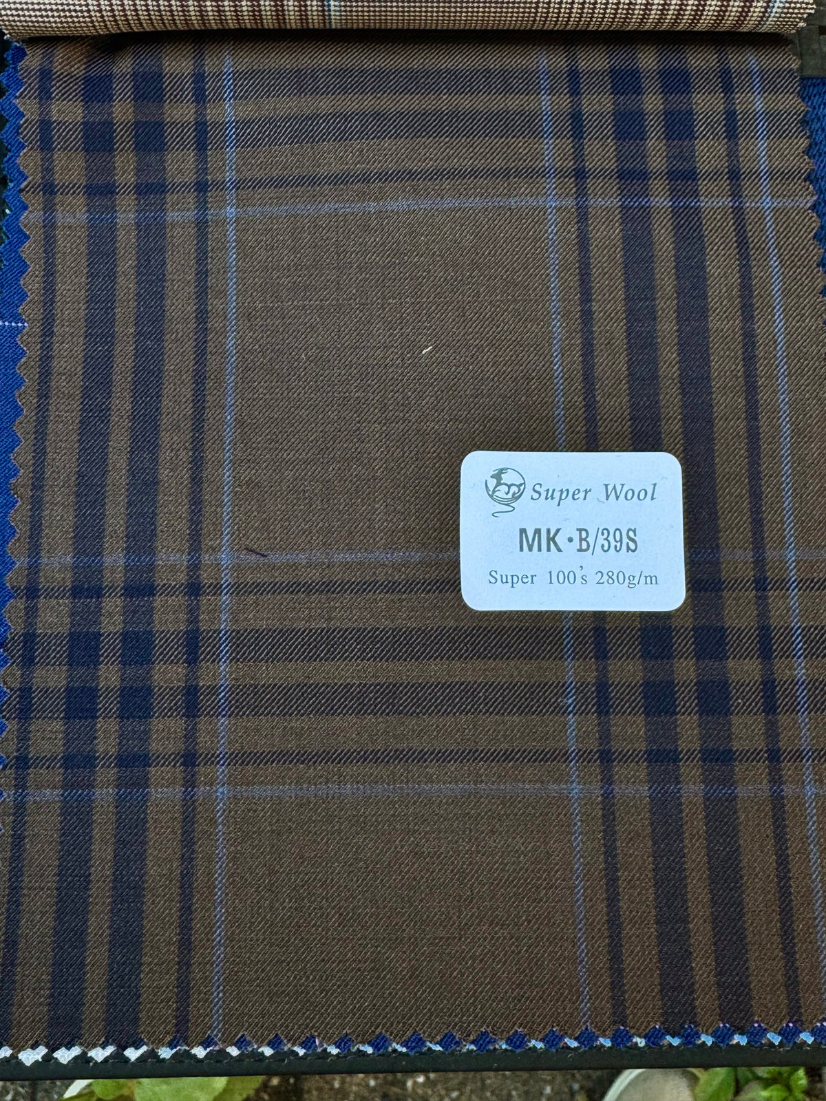 A close-up of a brown plaid fabric swatch with navy and blue lines, featuring a white label in the center that reads 'Super Wool MK-B/39S Super 100's 280g/m'.